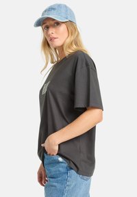 Camiseta de algodón gris con un corte holgado, mangas cortas, y un bolsillo con un diseño estampado, combinada con una gorra de denim azul claro.