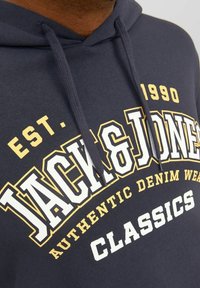 Marineblauer Hoodie mit Grafikdruck. Mit weißem und gelbem Text: "JACK & JONES", "AUTHENTIC DENIM WEAR" und "CLASSICS". Kapuze mit Zugband.