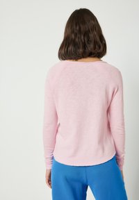 Žena s hnědými vlasy po ramena, oblečená v světle růžovém tričku s dlouhým rukávem a jasně modrých kalhotách, viděná zezadu na jednobarevném pozadí.