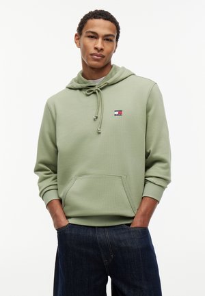 Νέος άνδρας με ανοιχτοπράσινο φούτερ με κουκούλα και λογότυπο στο στήθος και σκούρο μπλε τζιν, στέκεται με τα χέρια στις τσέπες σε λευκό φόντο.