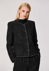Veste en tweed noire avec une surface texturée, un col rond et des boutons proéminents. Elle présente des coutures blanches sur les bords et des épaules structurées.
