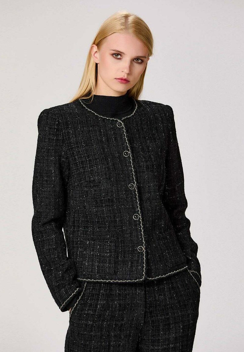 Veste en tweed noire avec une surface texturée, un col rond et des boutons proéminents. Elle présente des coutures blanches sur les bords et des épaules structurées.