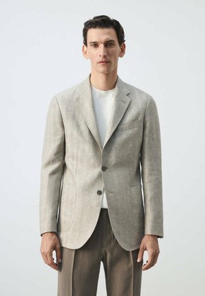 Homme portant un blazer beige sur une chemise blanche et un pantalon marron, debout devant un fond clair uni, face à l’avant.