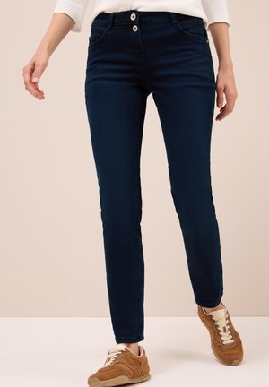 Jeans in denim blu navy con vestibilità slim, dotati di due chiusure a bottone e tasche frontali. Indossati con sneaker marroni su uno sfondo chiaro.
