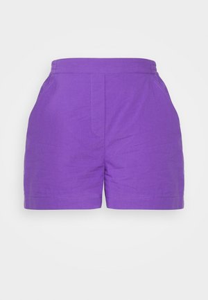 Henkilö, jolla on päällään valkoinen pitkähihainen pitsipaita, violettit korkeavyötäröiset shortsit ja valkoiset korkokengät, seisoo tavallista seinää vasten.