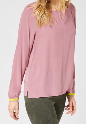 Kvinde iført en langærmet støvet lyserød bluse med gule stribede manchetter og olivengrønne bukser, stående mod en ensfarvet lys baggrund.