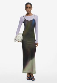 Langes, gradient Kleid in dunklem Grün und Braun, über einem helllila Langarmoberteil. Eine weiße Tasche und blaue Flip-Flops ergänzen den Look.