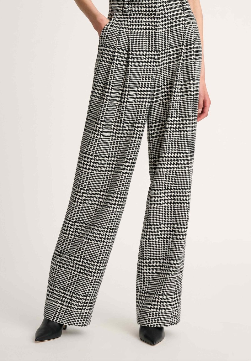 Pantalon large à motif pied-de-poule noir et blanc porté avec des bottes noires à bout pointu, mains dans les poches.
