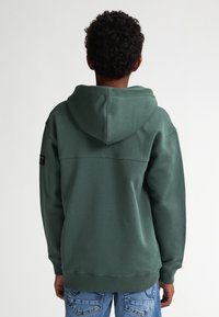 Groene hoodie met een eenvoudig ontwerp, voorzien van geribbelde mouwen, een kangoeroezak en een label op de linkermouw.