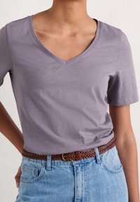 Persona che indossa una maglietta a V di colore lilla chiaro infilata in jeans a vita alta di colore blu chiaro con una cintura intrecciata marrone.