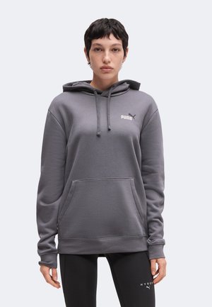 Persona con capelli corti e scuri indossa una felpa grigia Puma e leggings neri, in piedi rivolta in avanti su uno sfondo chiaro.