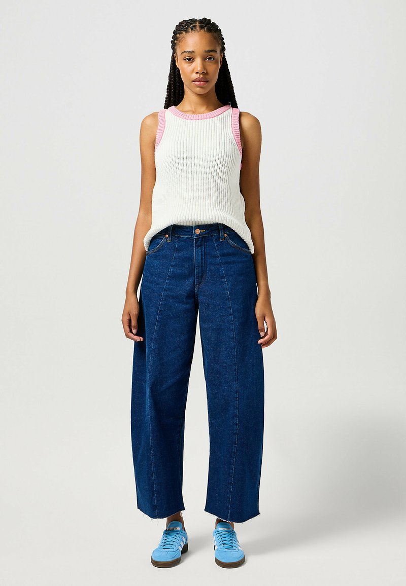 Débardeur blanc à rayures avec un contour rose, associé à un jean large bleu foncé, coupé au-dessus de la cheville, et des baskets bleues.