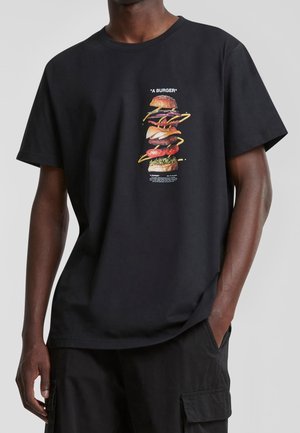 Mężczyzna w czarnej koszulce z grafiką rozłożonych składników burgera i napisem „A BURGER” na środku przodu.