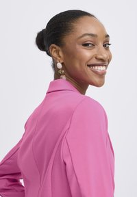 Pinkfarbener Blazer mit langen Ärmeln und taillierter Passform, aus weichem Stoff und mit dezenten Schulterdetails. Schmuckohrringe mit pastellfarbenen Steinen.