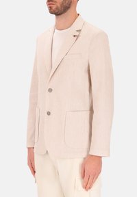 Blazer a righe beige con due bottoni, una tasca sul petto e due tasche frontali. Realizzato in tessuto testurizzato, presenta una leggera lucentezza.