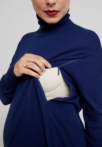 Navyblauer Rollkragenpullover mit einem Reißverschluss, der einen hellen BH darunter zeigt. Der Stoff wirkt weich und strukturiert mit einer lockeren Passform.