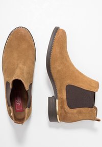 Un paio di stivali Chelsea in suede beige con pannelli elastici neri ai lati e suole marrone scuro, mostrati da vista superiore e laterale.