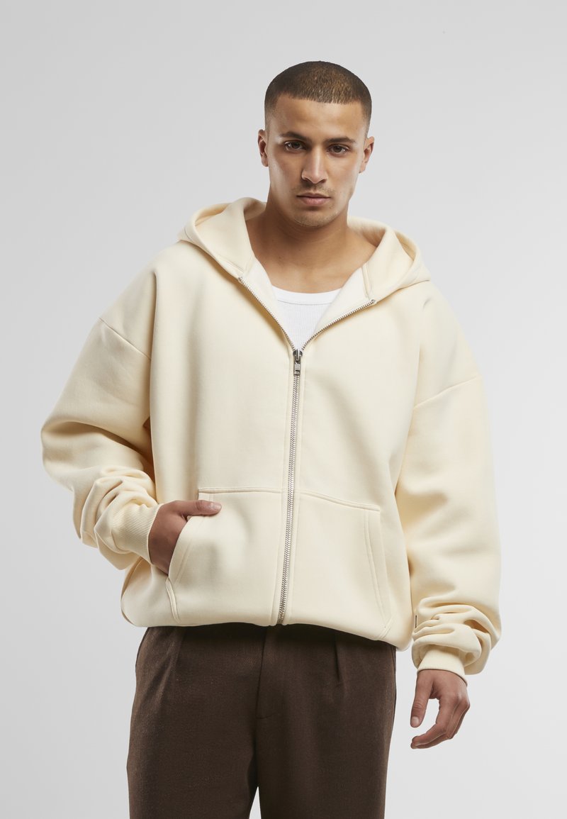 Hellbeige Zip-Hoodie aus weichem Stoff, mit einer großen Kapuze, frontalen Taschen und entspannter Passform. Kombiniert mit braunen Hosen.