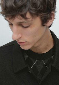 Schwarze Jacke mit Kragen über einem dunklen Pullover mit Rautenmuster. Lockiges Haar und neutraler Hautton, der sich auf die Details des Hemdes konzentriert. Weiches, strukturiertes Aussehen.