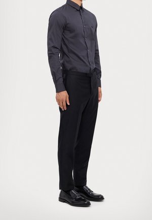 Man draagt een aansluitend donkergrijs overhemd met knopen, zwarte nette broek en zwarte leren loafers, staand tegen een eenvoudige lichte achtergrond.