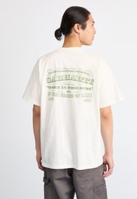 Hombre con cabello oscuro recogido en un moño, viste una camiseta crema de Carhartt con texto verde en la parte de atrás y pantalones cargo grises, de espaldas a la cámara.