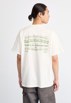 Hombre con cabello oscuro recogido en un moño, viste una camiseta crema de Carhartt con texto verde en la parte de atrás y pantalones cargo grises, de espaldas a la cámara.