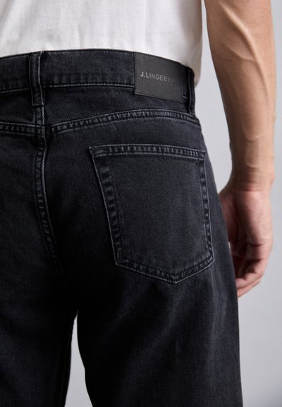 Black denim jeans with a back pocket detail, featuring a textured label at the waistband. The fabric shows a slight stretch and a smooth finish.