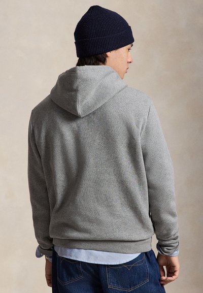 Un homme portant un bonnet en maille bleu marine, un sweat-shirt gris à capuche et un jean se tient dos à l’appareil photo et regarde par-dessus son épaule.