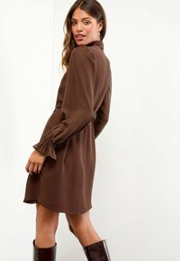 Braunes langärmeliges Kleid mit tailliertem Schnitt, gerafften Manschetten und einem Kragen, gefertigt aus glattem Stoff. Kombiniert mit knielangen, glänzenden schwarzen Stiefeln.