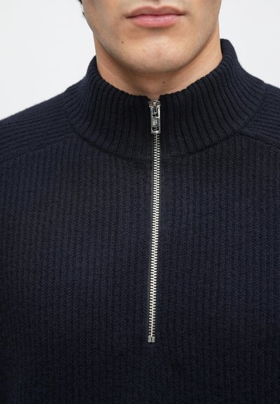 Navy ribbed knit sweater with a high collar and silver zipper closure. Textured fabric and fitted design details are visible.
