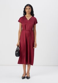 Robe midi bordeaux en tissu lisse avec des manches courtes, un col en V, une taille cintrée et une ceinture ; associée à un sac à main noir et des escarpins pointus.