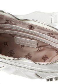 Otevřená bílá kožená kabelka s béžovým interiérem, který je potažen vzorem log „Steve Madden“ a obsahuje vnitřní kapsu na zip s logem značky.