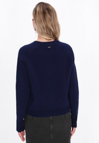 Pulover tricotat de culoare bleumarin, cu guler rotund și mâneci raglan; are un tiv striat și un detaliu subtil al logo-ului la gulerul din spate.