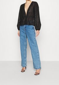 Blouse noire, en dentelle transparente, avec des manches longues et un décolleté en V profond ; associée à un jean droit bleu clair et des chaussures à talons noires à lanières.