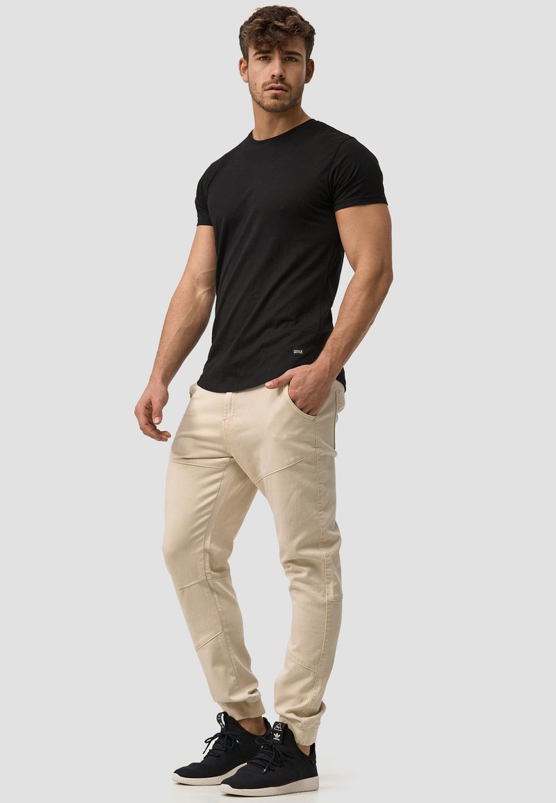 T-shirt noir à manches courtes associé à un pantalon de jogging beige avec un design texturé, et des chaussures de sport noires. Tenue simple et décontractée.