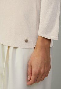 Hellrose Strickoberteil mit Dreiviertelärmeln und texturiertem Finish, mit einem silbernen Logoakzent am unteren Saum. Weiße Hosen sind teilweise sichtbar.