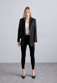 Zwarte blazer met twee knopen, witte cropped top, zwarte legging met ritsen en puntige zwarte hakken; model houdt een kleine zwarte tas vast.