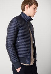 Veste rembourrée marine avec un quilt horizontal, col haut, fermeture éclair et poignets côtelés. Dispose de deux poches latérales. Matériau lisse et léger.