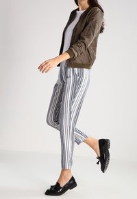 Olivgrön bomberjacka med ribbade muddar, vit T-shirt och svarta och vita randiga tapered-byxor; svarta skor med en rosettaccent.