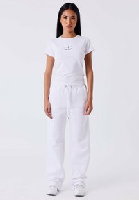 Femme aux longs cheveux noirs portant un t-shirt blanc avec logo, un pantalon de survêtement blanc ample et des baskets blanches, se tenant devant un fond blanc uni.