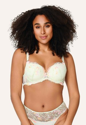 Žena kučeravými vlasmi v zladenom svetlozelenom kvetinovom čipkovanom podprsenkovom a nohavičkovom sete, stojaca pred jednoduchým svetlým pozadím.