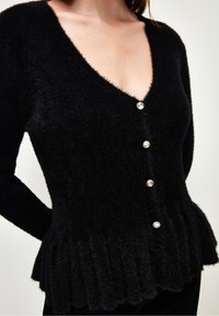 Czarny dziany kardigan z dekoltem w kształcie litery V, pięcioma błyszczącymi guzikami oraz fakturowanym dołem w stylu peplum. Materiał jest miękki i puszysty.