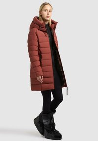 Giacca a vento in ruggine, con colletto alto, chiusura con zip e tasche laterali. Abbinata a stivali neri imbottiti e leggings.