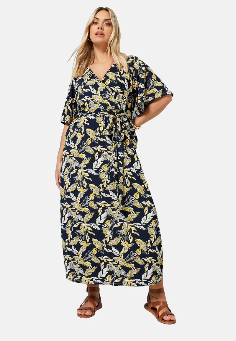 Maxiklänning med marinblå bakgrund och vita samt gula bladmönster, v-ringad halslinje, korta kimonoärmar och midjeband. Bruna sandaler.