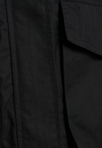 Primo piano di tessuto nero con texture liscia, cuciture visibili e design con tasca a patta. Il materiale sembra leggero e opaco.