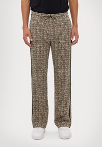 Kuosi Housut beigen ja mustan geometrisen kuvion kanssa, nyöri vyötäröllä, mustat sivuviivat ja hengittävä kangas. Valkoiset lenkkarit jalassa.