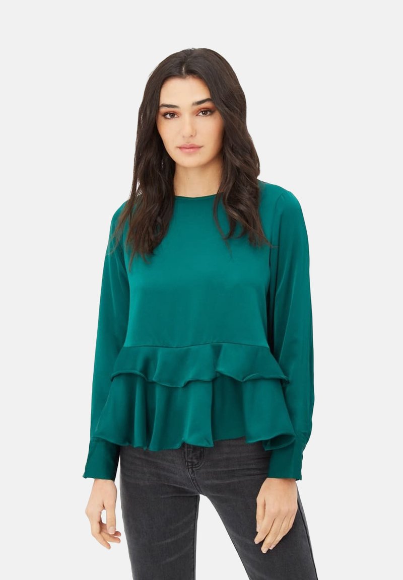 Blusa de manga larga color verde azulado con acabado satinado, que presenta un dobladillo en capas con volantes y un cuello redondo, combinada con jeans oscuros.