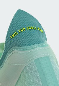 Teal sneakers med teksturert overdel i lett materiale, og gult brodert tekst "THIS TOO SHALL PASS" på hælen.