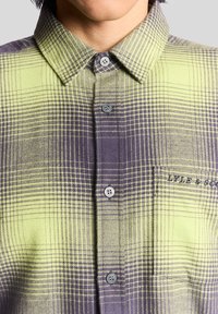 Kurzärmliges, kariertes Hemd in grünen und lila Tönen, mit einem Knopfkragen, vier Knöpfen und einem kleinen Logo auf der linken Seite.