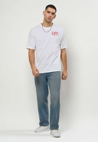 Νεαρός άνδρας στέκεται φορώντας λευκό t-shirt Kappa, μπλε τζιν, λευκά αθλητικά παπούτσια και ασημένιο μενταγιόν σε απλό ανοιχτό φόντο.